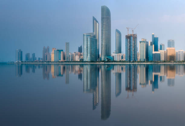 View of Abu Dhabi Skyline at sunrise with cloudy sky and reflection of buildings on water, United Arab Emirates